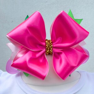 May include: A large, bright pink satin bow with a gold-colored center, layered over a white shirt. The shirt has the words "Melody berry" in green script. Green and purple accents are visible.