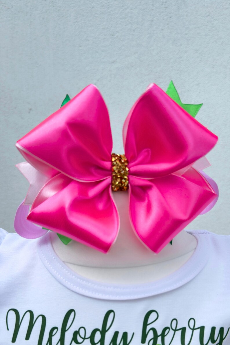 May include: A large, bright pink satin bow with a gold-colored center, layered over a white shirt. The shirt has the words "Melody berry" in green script. Green and purple accents are visible.