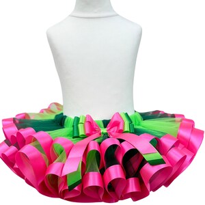 May include: A vibrant tutu skirt in shades of pink, green, and lime green, displayed on a white mannequin. The skirt features layers of tulle and satin ribbons, with a large bow at the waist. The design is eye-catching and suitable for dance or dress-up.