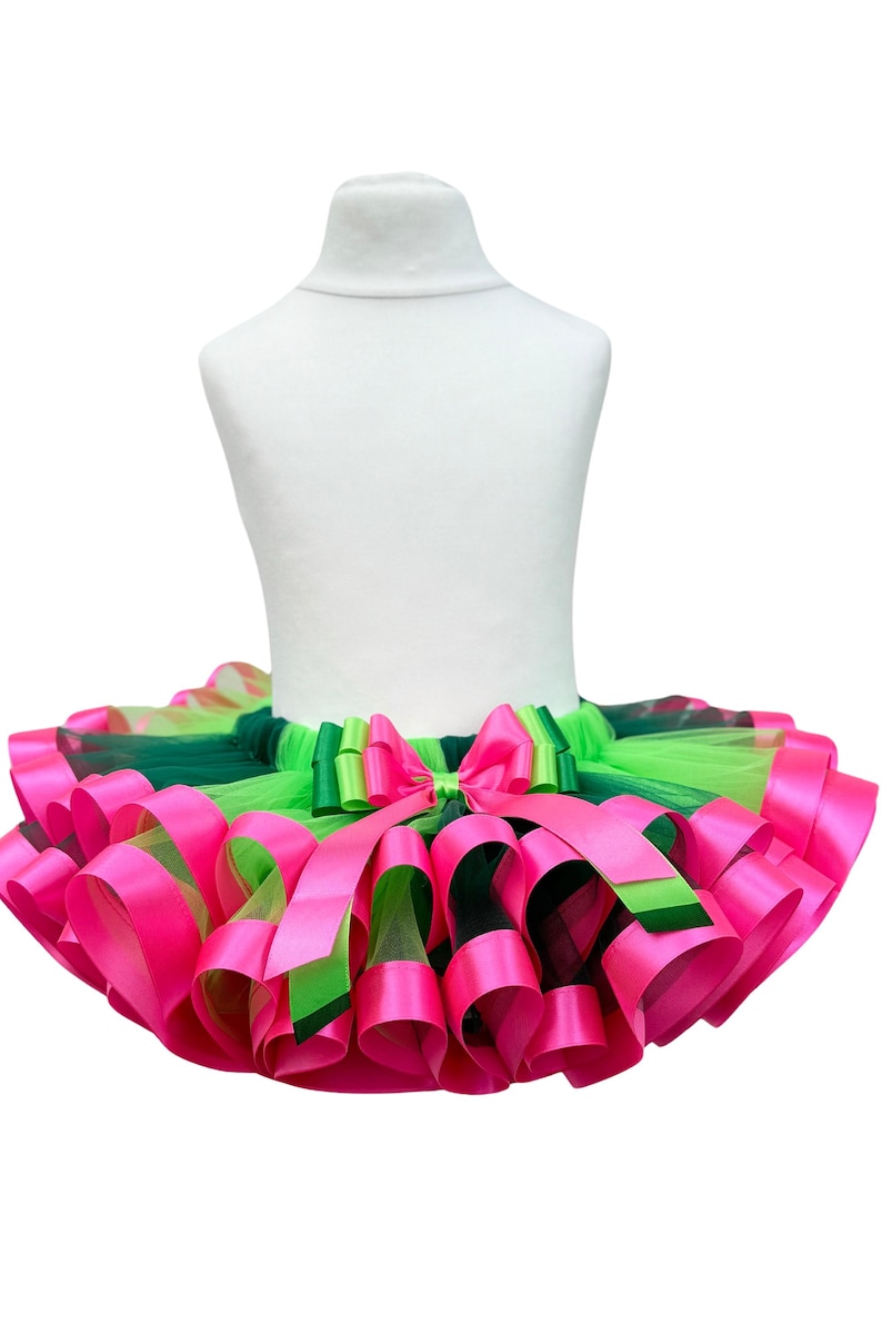 May include: A vibrant tutu skirt in shades of pink, green, and lime green, displayed on a white mannequin. The skirt features layers of tulle and satin ribbons, with a large bow at the waist. The design is eye-catching and suitable for dance or dress-up.