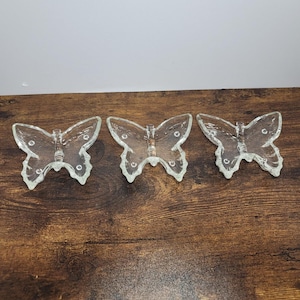 May include: Three clear glass dishes shaped like butterflies. Each dish features a textured surface with raised edges and a central body section. The glass is transparent and reflects light, showing a detailed wing pattern with small circular indentations.