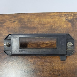 May include: A black rectangular plastic automotive license plate light housing featuring a central rectangular opening, two metal mounting studs on the sides, and the molded text F87F 12B523 PW >PP-GF 10< on the front surface.