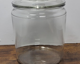 May include: A clear glass storage jar with a cylindrical body and a matching glass lid featuring a central knob handle. The container has a wide mouth and a smooth, transparent surface, providing a full view of the interior space.