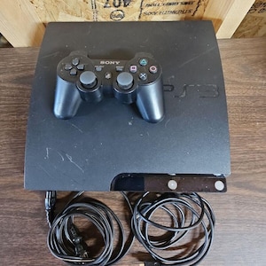 May include: A black Sony PlayStation 3 game console with a matching black wireless controller resting on top. The console features the PS3 logo embossed on the surface. Two black power and charging cables are coiled in front of the console unit.