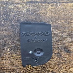 May include: A black Traxxas Link wireless module featuring a rectangular shape with rounded corners. The front surface displays the embossed Traxxas Link logo, a small circular indicator light, a central button, and the text PATENT TRAXXAS.COM/PAT.