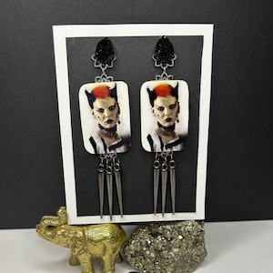 May include: A pair of dangle earrings featuring a black and white image of a woman with red hair. The earrings have a silver spike design and black accents.