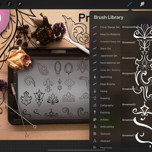 May include: A digital drawing tablet with a stylus and a selection of 20 decorative stamps displayed on the screen. The stamps are black and white and feature various floral and geometric designs. The tablet is on a wooden surface with a tattoo machine and other tools.