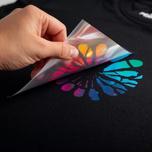 Pode incluir: Uma camiseta preta com um design colorido e iridescente sendo removido. O design apresenta um padrão floral com tons de amarelo, laranja, vermelho, azul e verde. A imagem também inclui o texto "YOU PERSONALISE".