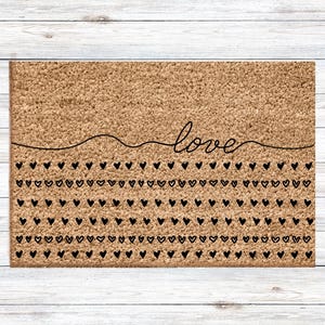 Peut inclure: Un paillasson rectangulaire en fibre de coco naturelle. Le mot "love" est écrit en cursive au-dessus de rangées de cœurs noirs. Le texte "Choisissez le mot de votre choix dans le menu des modèles" est à gauche.