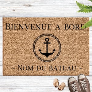 Peut inclure: Un paillasson rectangulaire avec le texte "BIENVENUE A BORD" et "- NOM DU BATEAU -" en noir. Une ancre dans un cercle de corde est au centre. Le tapis est marron et repose sur une surface en bois blanc. Des chaussures et des baskets sont en bas.