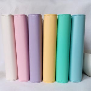 Op de afbeelding: Een rij van zes pastelkleurige agenda's. De agenda's zijn naast elkaar geplaatst, elk in een andere kleur: crème, roze, lavendel, geel, blauwgroen en lichtblauw. De covers zijn glad en de ruggen zijn gestikt.