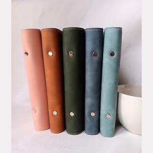 Puede incluir: Una fila de cinco diarios de cuero en colores pastel: rosa, marrón, verde oscuro, azul y turquesa. Cada diario tiene un cierre de botón plateado y un lomo redondeado con costuras. Una taza de cerámica blanca es parcialmente visible.