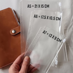 Può includere: Un'agenda in pelle marrone accanto a buste trasparenti in plastica per raccoglitori nei formati A5 (21 x 15 cm), A6 (17,5 x 10,5 cm) e A7 (13 x 9 cm). Ogni busta ha una chiusura a zip ed è progettata per raccoglitori ad anelli. Le dimensioni sono stampate.