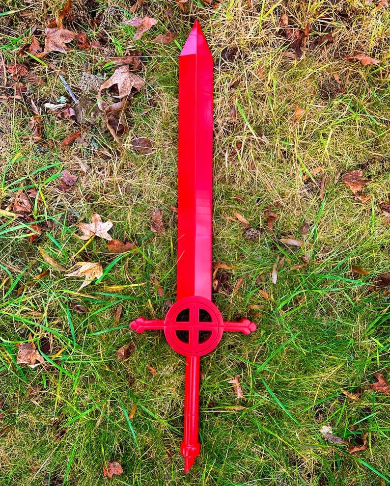 May include: A red plastic sword with a circular cross guard. The sword is lying on a bed of green grass.