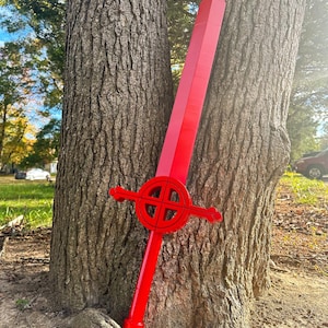 May include: A bright red sword with a circular guard and a wooden handle. The sword is leaning against two tree trunks.