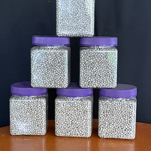 May include: Six clear plastic jars with purple lids filled with small white granules. The jars are arranged in a pyramid shape on a wooden surface.