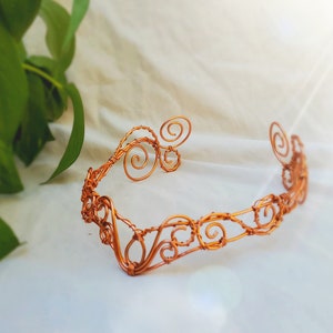 May include: A copper wire crown with a delicate, intricate design featuring swirls and loops. The crown is handmade and has a rustic, earthy aesthetic.