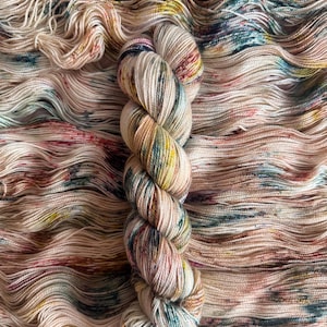 May include: A skein of hand-dyed yarn with a variegated color palette. The yarn is a creamy off-white base with splashes of pink, yellow, teal, and brown. The yarn is soft and appears to be a natural fiber, perfect for knitting or crocheting projects.
