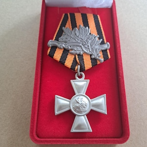 May include: A silver-colored medal in a red velvet box. The medal features a cross design with a central emblem and is suspended from a ribbon with black and orange stripes. A silver laurel wreath is attached to the ribbon.