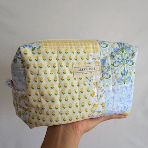 May include: A quilted fabric pouch featuring a patchwork design of yellow floral, yellow gingham, and blue floral patterns. A rectangular fabric label on the front displays the text GREEN ROSE HANDMADE in black ink on a cream-colored background.