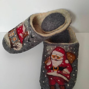 May include: A pair of gray felt slippers with a winter theme. One slipper features a house with a chimney and a snowman, while the other slipper features Santa Claus with a bag of gifts. Both slippers have white snowflakes on a gray background.