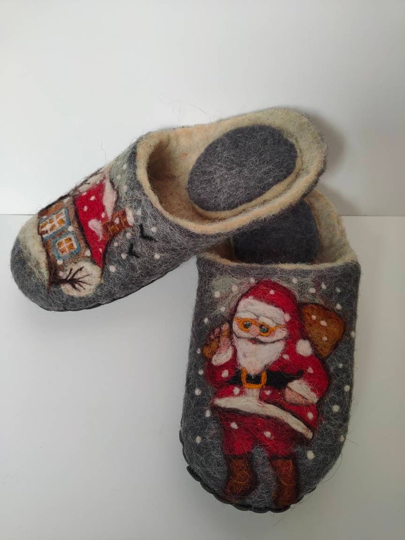 May include: A pair of gray felt slippers with a winter theme. One slipper features a house with a chimney and a snowman, while the other slipper features Santa Claus with a bag of gifts. Both slippers have white snowflakes on a gray background.