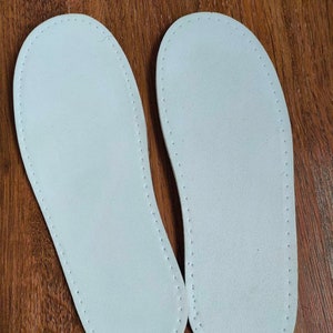 May include: Two white leather insoles with a stitched edge. The insoles are on a wooden surface.