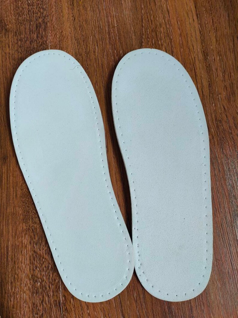 May include: Two white leather insoles with a stitched edge. The insoles are on a wooden surface.