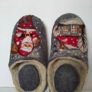 May include: A pair of gray felt slippers with a Christmas theme. One slipper features a Santa Claus design, while the other has a house with snow-covered trees.
