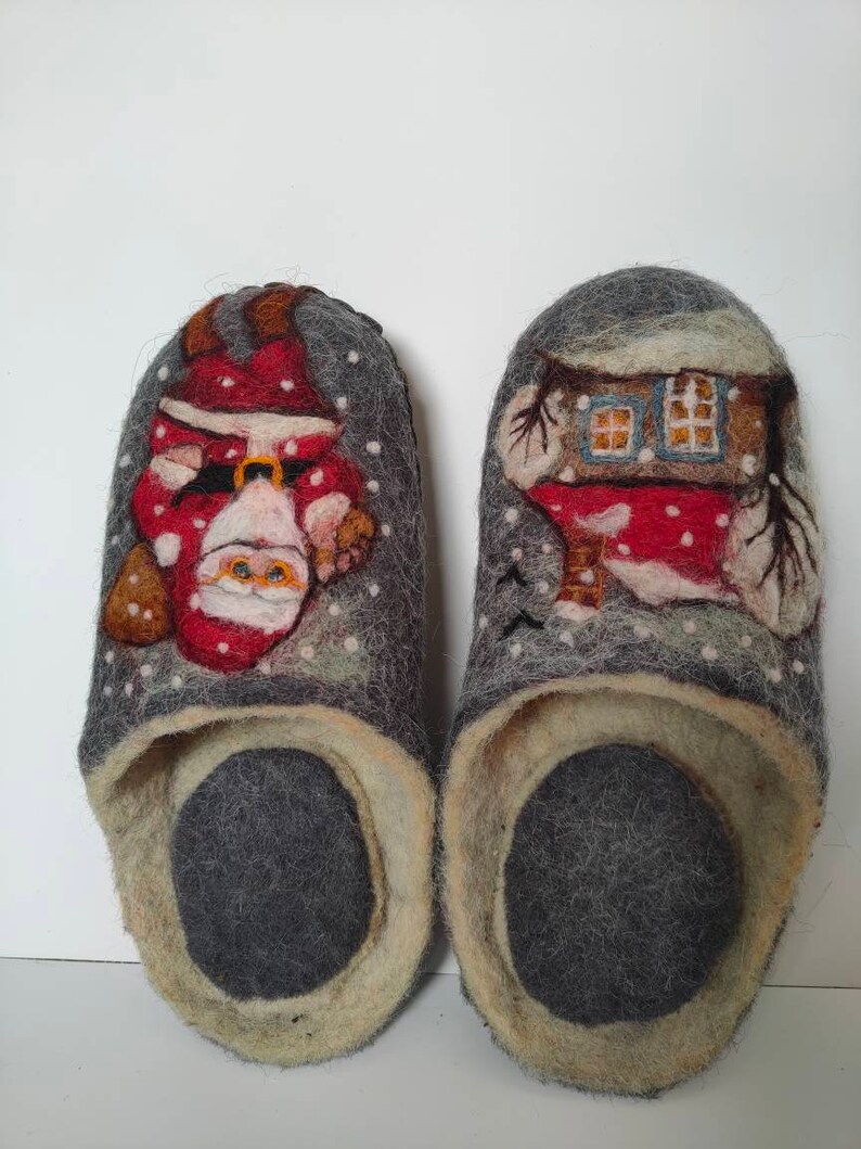 May include: A pair of gray felt slippers with a Christmas theme. One slipper features a Santa Claus design, while the other has a house with snow-covered trees.