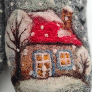 May include: A pair of gray felt slippers with a winter scene design. The scene features a small house with a red roof and white snow, a brown chimney, and two windows with blue frames. The house is surrounded by white snow and a few trees.