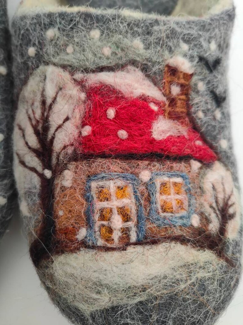 May include: A pair of gray felt slippers with a winter scene design. The scene features a small house with a red roof and white snow, a brown chimney, and two windows with blue frames. The house is surrounded by white snow and a few trees.