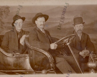May include: Three men in hats are sitting in a vintage car. The man in the middle is driving and holding the steering wheel. The man on the left is sitting in the passenger seat. The man on the right is sitting in the back seat. The car is a dark color and has a wooden dashboard.