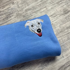 K&ouml;nnte beinhalten: Ein hellblauer Pullover mit einem gestickten Bild eines Hundefaces. Der Hund hat wei&szlig;es Fell und eine rote Zunge.
