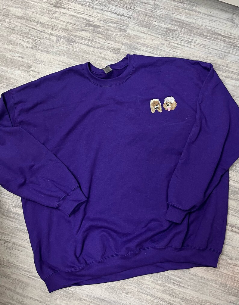 K&ouml;nnte beinhalten: Ein lilafarbener Pullover mit zwei gestickten Bildern von Hunden auf der Vorderseite. Die Hunde sind ein brauner und wei&szlig;er Pudel.