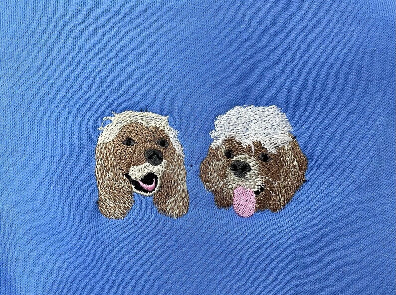 K&ouml;nnte beinhalten: Gestickte Abbildung von zwei Hunden auf blauem Hintergrund. Die Hunde sind braun mit wei&szlig;em Fell und schauen nach rechts.