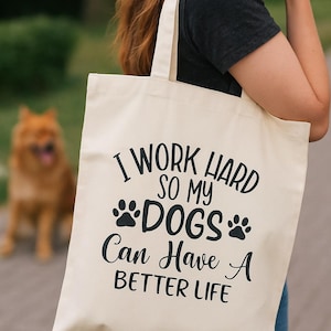 Könnte beinhalten: Cremefarbene Tragetasche mit schwarzem Text: „I WORK HARD SO MY DOGS CAN HAVE A BETTER LIFE“. Die Tasche wird von einer Person gehalten. Im Hintergrund ist ein Hund zu sehen. Die Tasche hat zwei Henkel.
