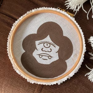 May include: A brown and white embroidery hoop featuring a one-eyed monster design. The monster has a large eye, a small nose, and a wide mouth with fangs.