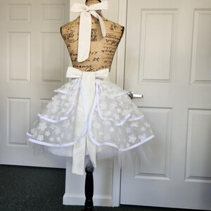 May include: A white apron featuring a layered tulle skirt adorned with small floral appliqu&eacute;s and white ribbon trim. The apron includes a wide white fabric waistband with a large bow at the back and a matching neck tie, displayed on a dress form.