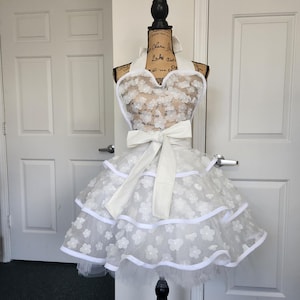 May include: A white apron featuring a sweetheart neckline and a tiered skirt. The apron is decorated with numerous small white floral appliqu&eacute;s and finished with white trim. A large white fabric bow is tied at the waist, and the apron secures with a halter-style neck strap.