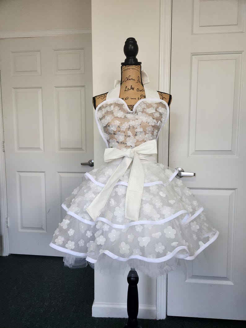 May include: A white apron featuring a sweetheart neckline and a tiered skirt. The apron is decorated with numerous small white floral appliqu&eacute;s and finished with white trim. A large white fabric bow is tied at the waist, and the apron secures with a halter-style neck strap.