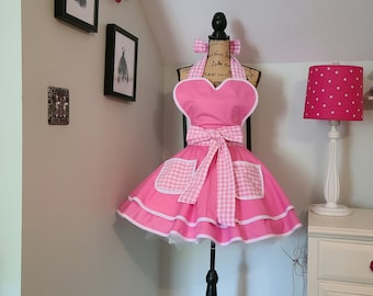 Op de afbeelding: Een roze en witte gingham schort met een hartvormig bovenstuk en een witte ruche. Het schort staat op een zwarte mannequinstandaard.