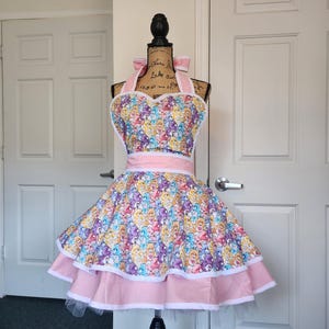 May include: A multi-tiered apron featuring a colorful Care Bears character print on a light background. The apron has a sweetheart neckline, a wide pink waistband, and a flared skirt with two layers of pink fabric trim and white piping edges.