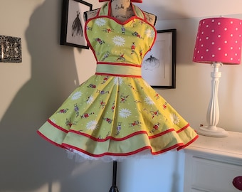 Op de afbeelding: Een groene en gele schort met een rode bies en een cartoonprint. De schort heeft een halternek-stijl en een volle rok. De schort staat op een mannequin.