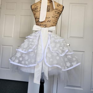 May include: A white apron featuring a tiered skirt with sheer floral lace overlays and solid white trim. The apron includes a wide waistband with a large bow at the back and a matching neck tie, displayed on a dress form with a vintage script print.
