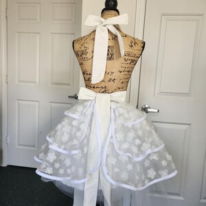May include: A white apron featuring a tiered skirt design with layered tulle fabric adorned with small white floral appliqu&eacute;s and solid white trim. The apron includes a wide white fabric tie at the waist and a matching tie at the neck.