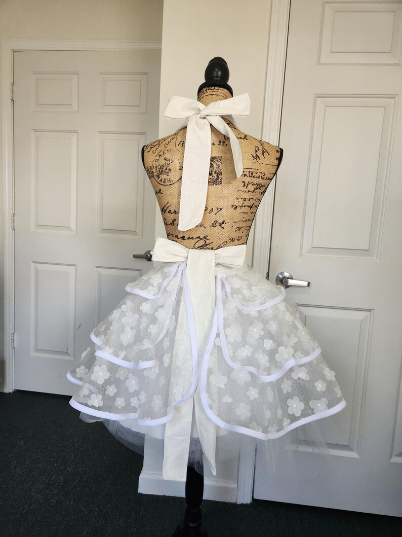 May include: A white apron featuring a tiered skirt design with layered tulle fabric adorned with small white floral appliqu&eacute;s and solid white trim. The apron includes a wide white fabric tie at the waist and a matching tie at the neck.
