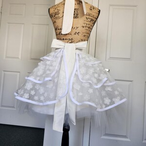 May include: A white apron featuring a tiered skirt with sheer floral lace overlays and solid white trim. The apron includes a wide waistband with a large bow at the back and a matching neck tie, displayed on a dress form with a vintage script print.