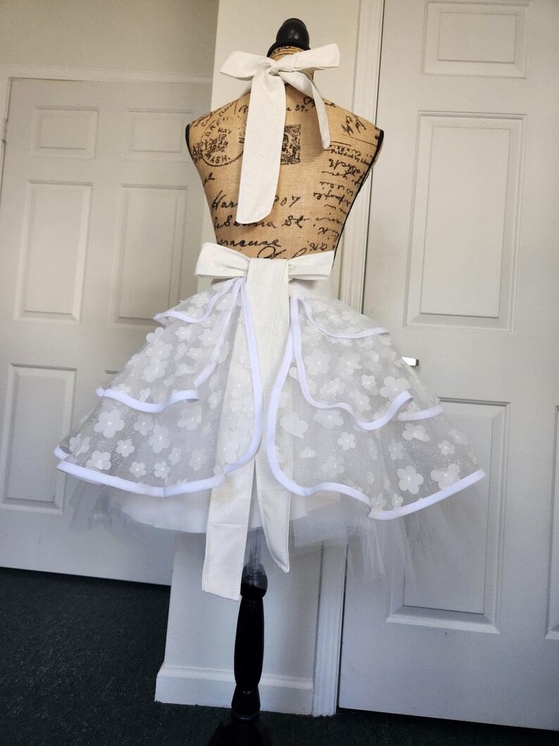 May include: A white apron featuring a tiered skirt with sheer floral lace overlays and solid white trim. The apron includes a wide waistband with a large bow at the back and a matching neck tie, displayed on a dress form with a vintage script print.
