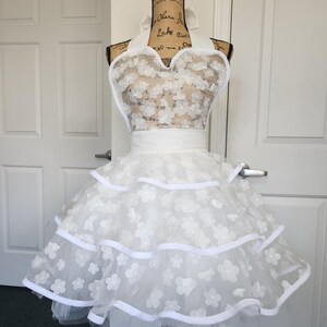 May include: A white apron featuring a halter neck design with a sweetheart neckline. The bodice and the three-tiered skirt are adorned with white floral appliqu&eacute;s and sheer mesh fabric, finished with solid white ribbon trim along the edges.